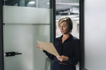Geschäftsfrau mit Headset, die am Eingang eines Büros ein Dokument liest - JOSEF17827