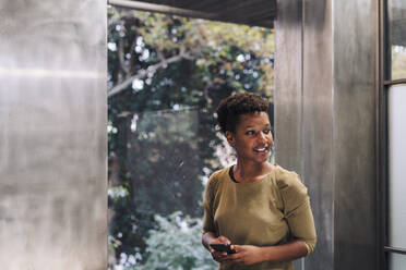 Happy businesswoman with smart phone standing by window - JOSEF17889