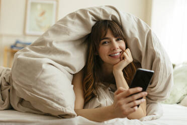 Happy woman with mobile phone wrapped in blanket at home - EBSF03013