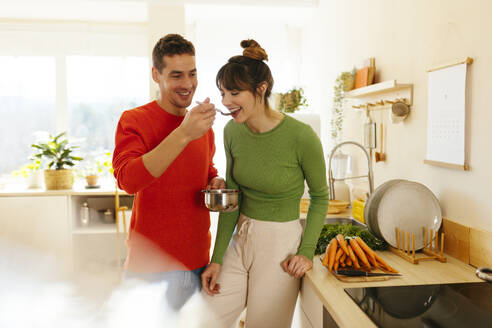 Happy man feeding woman standing in kitchen - EBSF03022