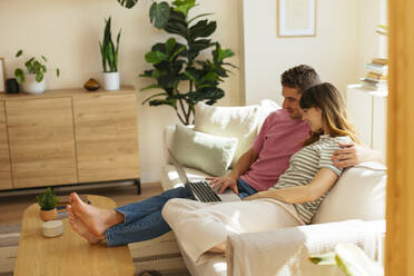 Boyfriend with arm around girlfriend using laptop on sofa in living room - EBSF03038