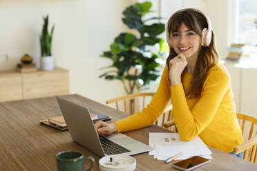 Glückliche Frau mit Kopfhörern, die mit einem Laptop am Tisch im Heimbüro sitzt - EBSF03051
