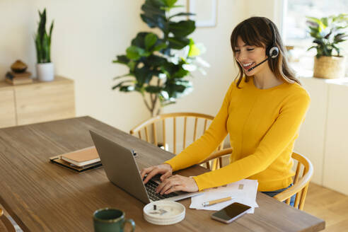 Happy freelancer wearing headset using laptop at home office - EBSF03058