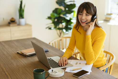 Happy freelancer talking on headset sitting with laptop at home office - EBSF03062