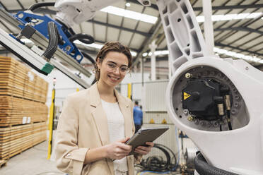 Lächelnder junger Ingenieur, der mit einem Tablet-PC vor einem Roboterarm steht - JCCMF09988