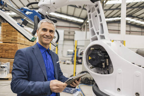 Lächelnder reifer Ingenieur mit Tablet-PC an einem Roboterarm - JCCMF09999