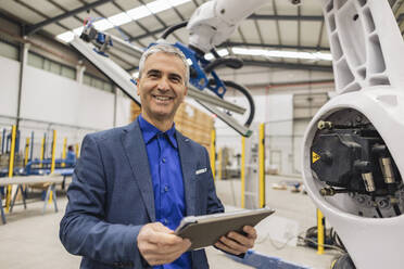 Glücklicher reifer Ingenieur mit Tablet-PC an einem Roboterarm - JCCMF10000