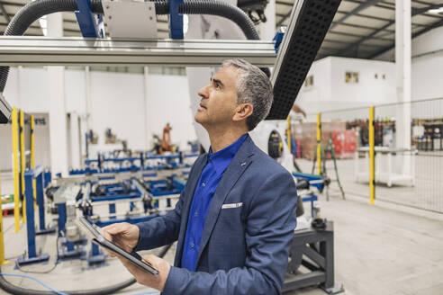 Älterer Geschäftsmann mit Tablet-PC, der einen Roboterarm in einer Fabrik untersucht - JCCMF10003