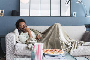 Lächelnder Mann mit Decke entspannt sich auf dem Sofa zu Hause - PNAF05117