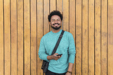 Smiling man with smart phone in front of wooden wall - PNAF05133