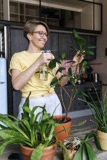 Smiling woman spraying water on plant leaves at home - VYF00956