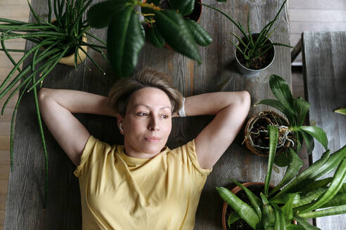 Nachdenkliche Frau mit den Händen hinter dem Kopf, die zu Hause inmitten von Pflanzen liegt - VYF00962