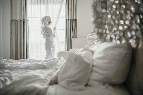 Woman standing near window in bedroom at home - MJRF00969