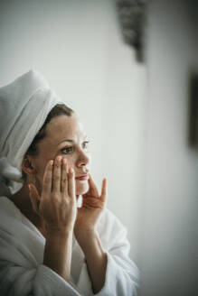 Frau trägt Feuchtigkeitscreme auf und schaut in den Spiegel zu Hause - MJRF00974