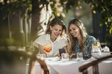 Glückliche Freunde, die sich ein Smartphone teilen, sitzen am Tisch im Restaurant - MJRF00984