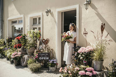 Lächelnde Frau mit Blumenstrauß in der Nähe der Tür eines Blumenladens - MJRF00994