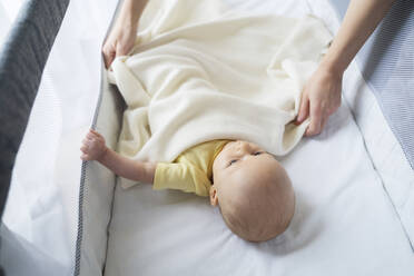 Mother putting blanket on son lying in crib - NJAF00298