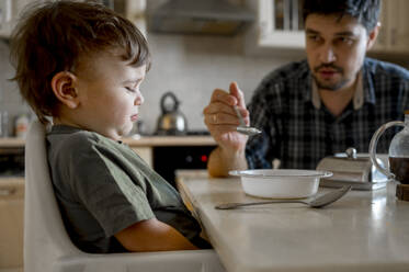 Vater füttert verärgerten Sohn am Esstisch sitzend im Haus - ANAF01123