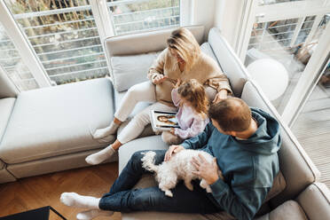 Tochter schaut sich Fotos mit Eltern und Hund an, die zu Hause auf dem Sofa sitzen - MOEF04453