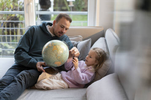 Father and daughter with globe sitting on sofa at home - MOEF04455