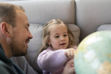 Daughter looking at globe with father on sofa at home - MOEF04456