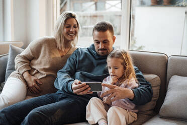 Parents sharing smart phone with daughter sitting on sofa at home - MOEF04468