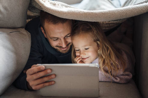 Vater und Tochter schauen auf einen Tablet-PC unter der Bettdecke - MOEF04486