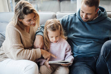 Daughter using tablet PC sitting with parents on sofa in living room - MOEF04495