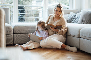 Mutter liest ein Buch, während ihre Tochter zu Hause einen Tablet-PC benutzt - MOEF04510