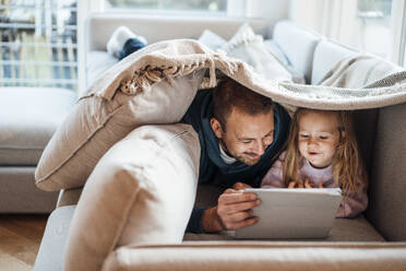 Happy father and daughter looking at tablet PC under blanket lying on sofa at home - MOEF04515