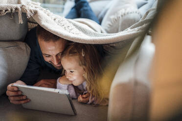 Happy man sharing tablet computer with daughter under blanket at home - MOEF04517