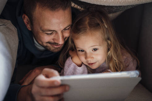 Vater und Tochter schauen sich zu Hause unter der Bettdecke einen Tablet-PC an - MOEF04518