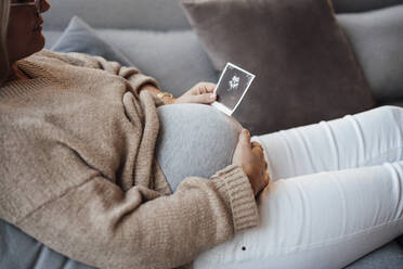 Schwangere Frau mit Ultraschallbild auf dem Sofa zu Hause - MOEF04523