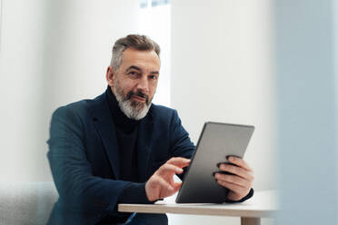 Smiling businessman sitting with tablet PC at desk - JOSEF17936