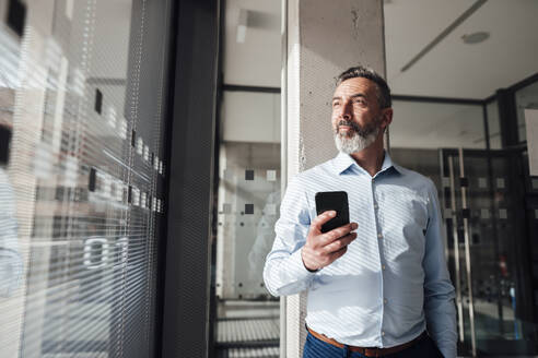 Nachdenklicher Geschäftsmann mit Smartphone im Büro stehend - JOSEF17949