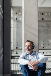 Thoughtful businessman listening to music through wireless headphones at office - JOSEF17959