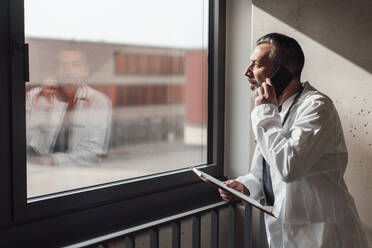 Arzt mit Klemmbrett im Gespräch mit Smartphone am Fenster - JOSEF17990