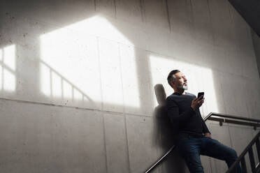 Thoughtful businessman standing with smart phone on staircase - JOSEF17996