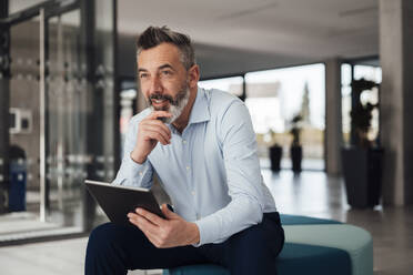 Thoughtful businessman sitting on seat with tablet PC at office - JOSEF18007