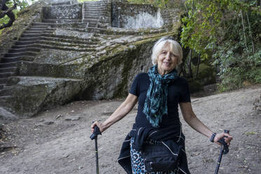 Lächelnde ältere Frau mit Wanderstöcken vor der etruskischen Pyramide von Bomarzo - MAMF02726