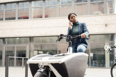 Geschäftsfrau, die auf einem Lastenfahrrad sitzend über ihr Smartphone spricht - JOSEF18042