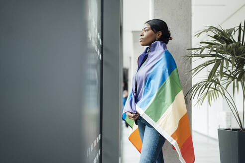 Junge Frau mit Regenbogenfahne, die im Büro aus dem Fenster schaut - JOSEF18086