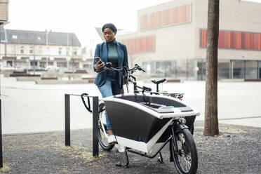 Geschäftsfrau surft im Internet über ihr Smartphone auf einem Lastenfahrrad sitzend - JOSEF18094