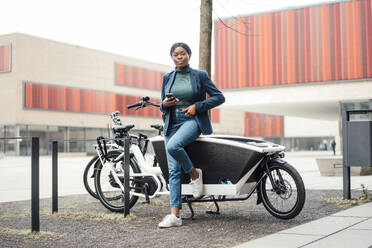 Confident businesswoman with smart phone leaning on cargo bike - JOSEF18096