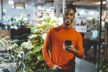 Happy young businessman holding mobile phone in office - JOSEF18110