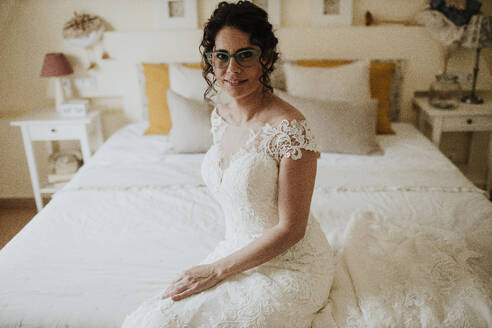 Lächelnde Braut im Hochzeitskleid auf dem Bett sitzend - GMLF01336