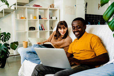 Glückliches multiethnisches Paar in Freizeitkleidung, das auf dem Sofa sitzt, ein Buch liest und einen Laptop im heimischen Wohnzimmer benutzt - ADSF43494