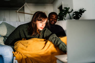 Junges gemischtrassiges Paar in Freizeitkleidung sitzt auf dem Sofa und surft auf dem Laptop, während es seine Freizeit zu Hause verbringt - ADSF43505