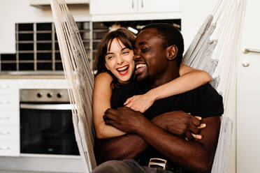 Fröhliches gemischtrassiges Paar in Freizeitkleidung, das sich umarmt und lacht, während es auf einem Schaukelstuhl in der Küche am Wochenende chillt - ADSF43506
