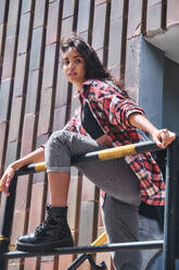 Side view of calm ethnic female model in colorful shirt and jeans with dark hair pondering and looking away while relaxing on stairway of building on street - ADSF43508